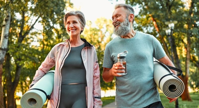 Two older adults walk outside with rolled yoga mats and a water bottle, smiling, in a park with trees.