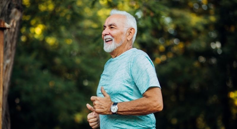 An older man with a white beard jogs outdoors in a light blue shirt, smiling, with trees in the background.