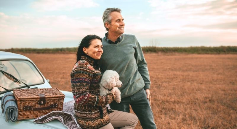 An older couple with a small white dog stands by a vintage car.