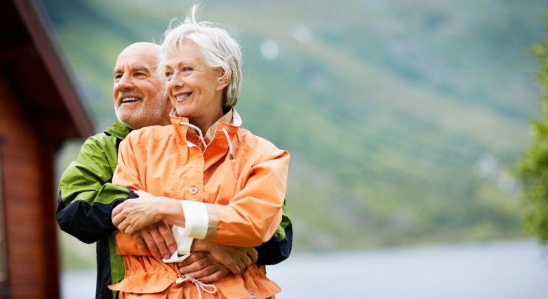 An older couple stands outdoors, embracing and smiling, with green hills and water blurred in the background.