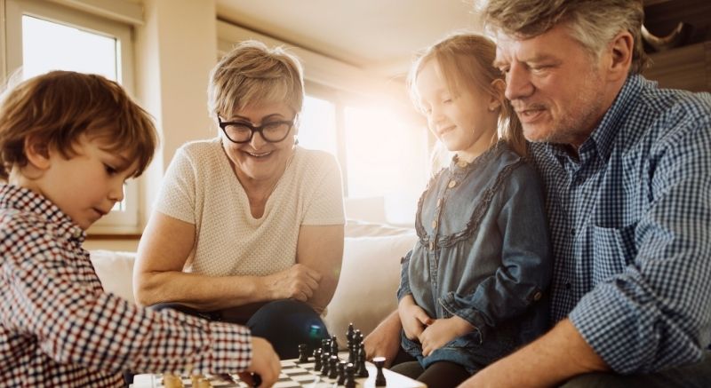 Two older adults and their grandchildren sit together playing chess on a table in a bright living room.