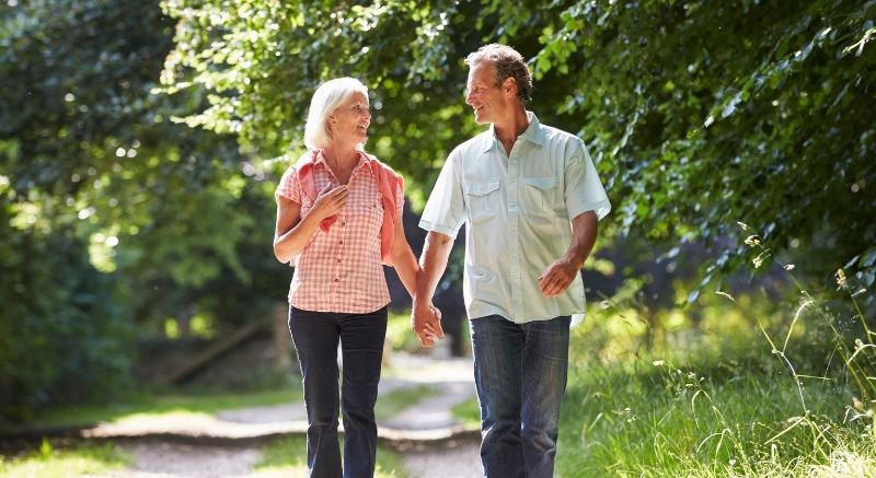 An older couple holding hands and walking together on a tree-lined path in a park on a sunny day.