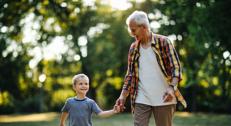 Older man and his grandson holding hands and walking outdoors in a park with trees in the background.