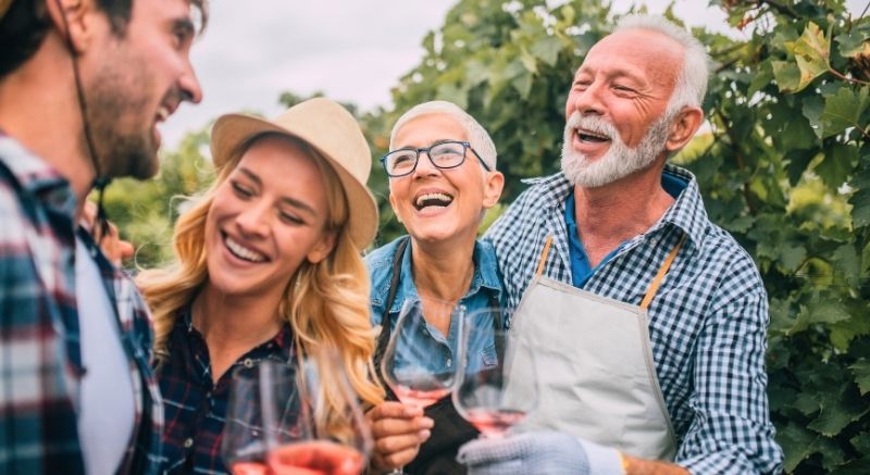 Four adults stand outdoors among grapevines, smiling and holding glasses of rosé wine, dressed in casual clothes.