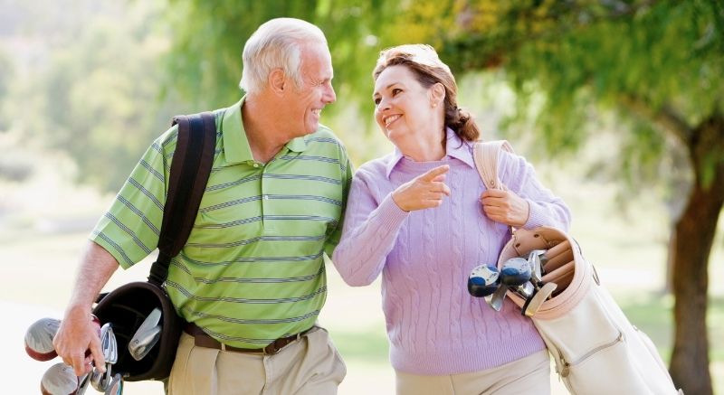 Older couple carrying golf bags and smiling at each other while walking on a golf course.