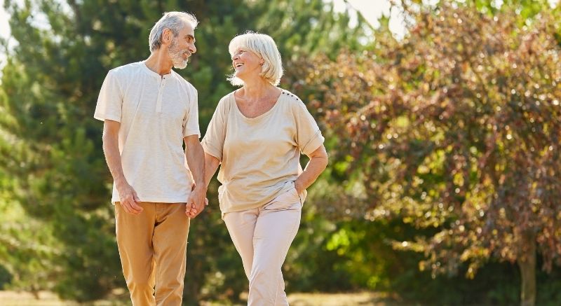 An older man and woman walk hand in hand outdoors, smiling, with trees and sunlight in the background.