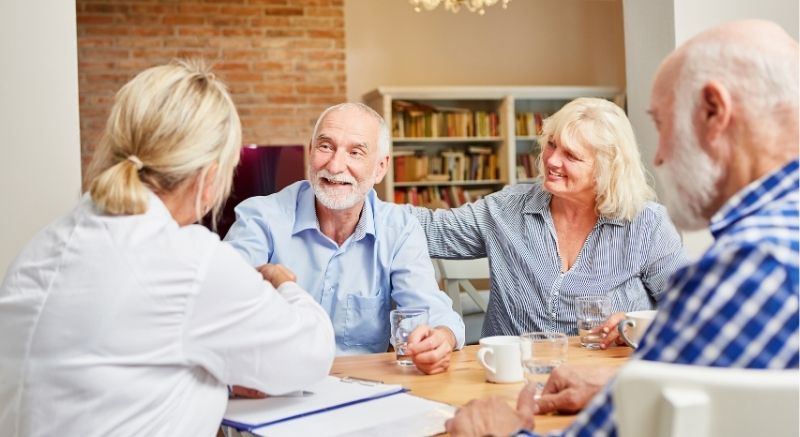 Four older adults discuss medicare supplements over drinks, seated at a table with bookshelves in the background.