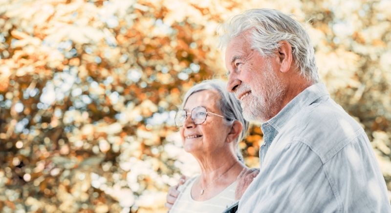 An older man and woman stand close together outdoors, smiling, with sunlight filtering through trees in the background.
