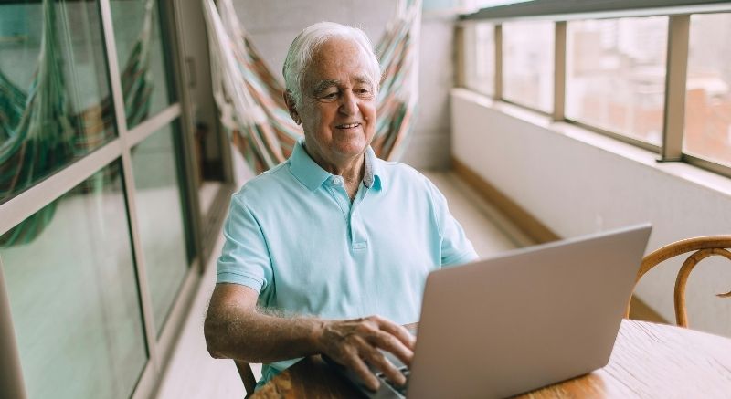 An older man in a light blue polo shirt uses his laptop for Medicare research while sitting on a balcony.