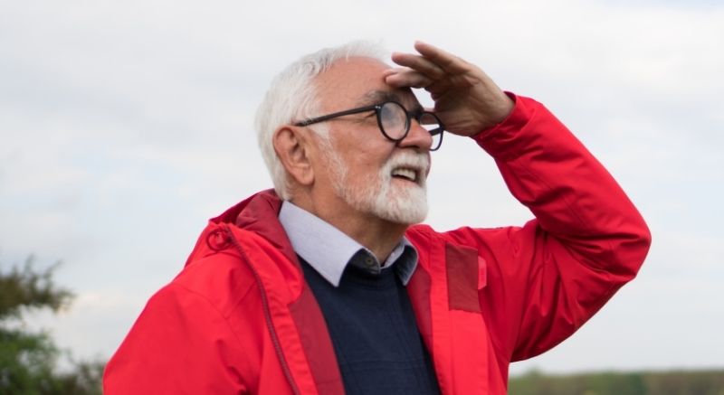 Older man in a red jacket looks into the distance with one hand raised to shade his eyes outdoors.