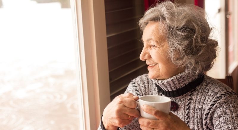 An older woman with gray hair holds a mug and looks out a window, wearing a gray sweater and smiling.