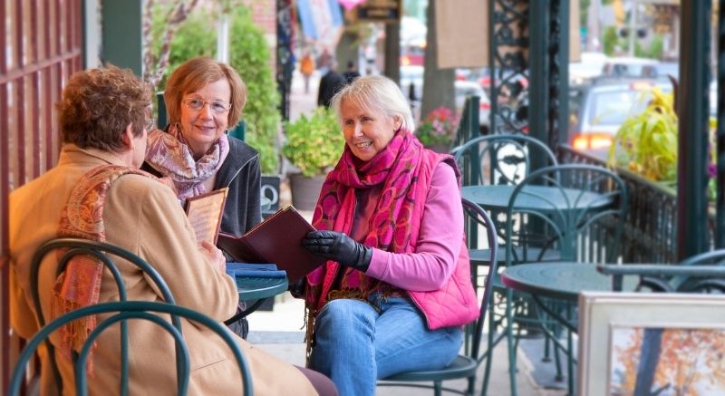 Three older women sit at an outdoor café table, talking and looking at menus on a chilly day.