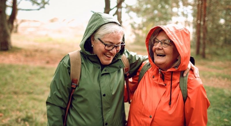 Two older adults in rain jackets and backpacks smile and stand outdoors among trees on a cloudy day.