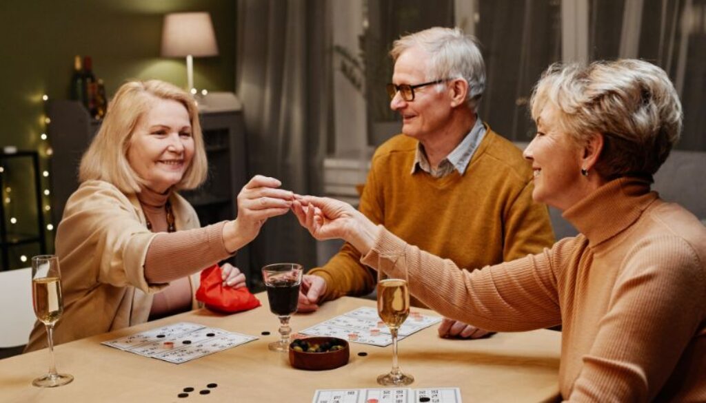 Three older adults, turning 65, sit at a table playing a board game with drinks and snacks in front of them.
