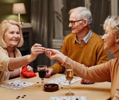 Three older adults, turning 65, sit at a table playing a board game with drinks and snacks in front of them.