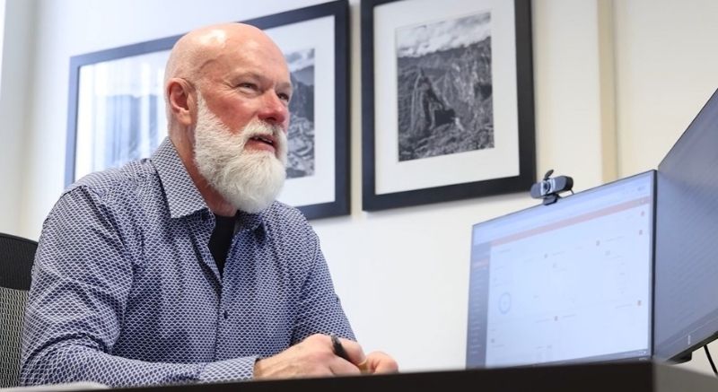 Bald man with a white beard sits at a desk, looking at a computer monitor in an office with framed pictures on the wall.