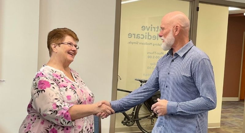 Two people are shaking hands indoors, both smiling. A bicycle is visible in the background near a glass door.