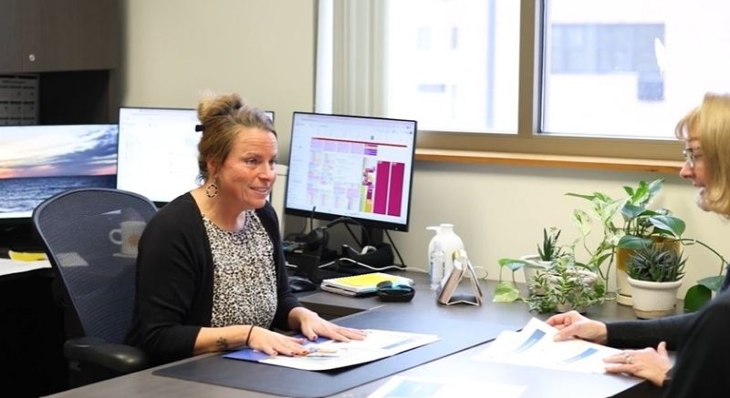 Two women sit across a desk in an office, discussing documents with computers and plants in the background.