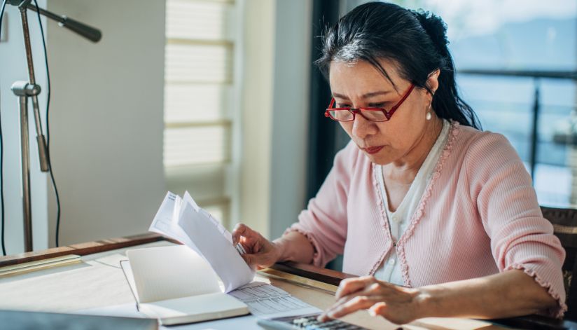 Woman in red glasses reviews papers about Medicare penalties at her desk, sunlight streaming through the window.