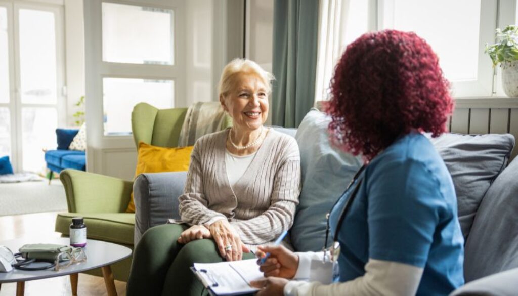 A healthcare professional talks with an older woman in a living room, taking notes on a clipboard.