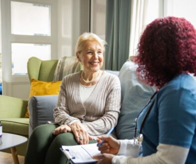 A healthcare professional talks with an older woman in a living room, taking notes on a clipboard.
