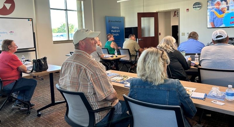 People sit at tables in a classroom setting, facing a presenter and a TV screen at the front of the room.