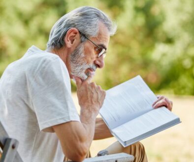 Older man with gray hair and glasses sits outdoors in a chair, reading a book and resting his chin on his hand.
