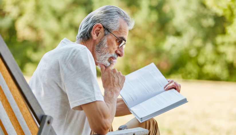 Older man with gray hair and glasses sits outdoors in a chair, reading a book and resting his chin on his hand.