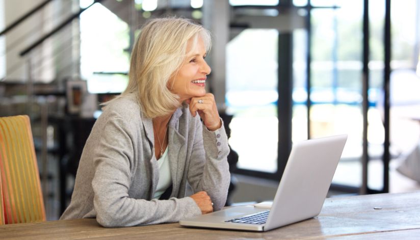 Older woman with gray hair smiles at her laptop, researching Medicare penalties in a bright, cheerful room.