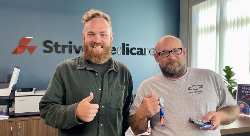 Two men stand indoors smiling and giving thumbs up in front of a wall sign reading Strive Medicare.