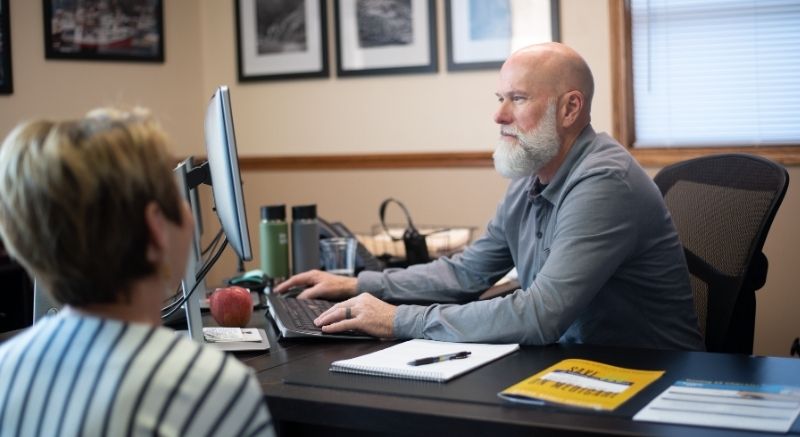 A man with a gray beard works at a computer while talking to a woman across a desk in an office setting.