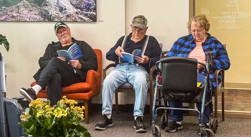 Three older adults sit in a waiting room reading pamphlets; one person uses a walker, and flowers are in the foreground.