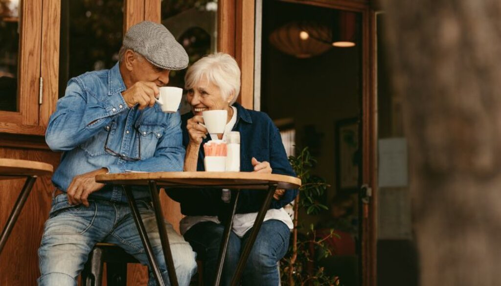 Two older adults sit at an outdoor café table, smiling and drinking coffee together.