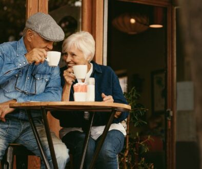 Two older adults sit at an outdoor café table, smiling and drinking coffee together.