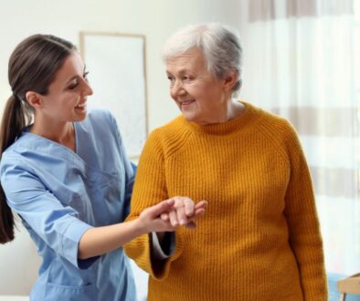 A young caregiver helps an older woman in a yellow sweater explore Medicare plan options in a bright, home-like room.