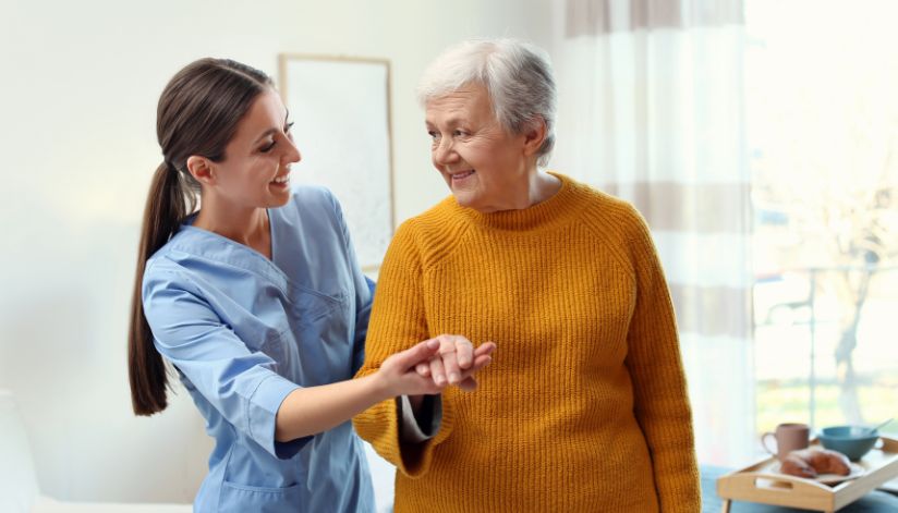 A young caregiver helps an older woman in a yellow sweater explore Medicare plan options in a bright, home-like room.