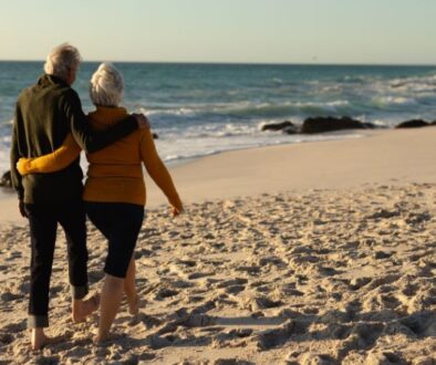 An older couple walks barefoot on a sandy beach, discussing Medicare plans with their arms around each other.