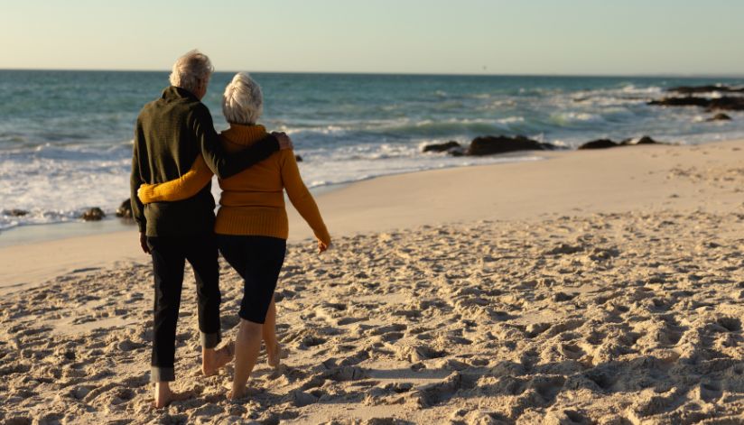 An older couple walks barefoot on a sandy beach, discussing Medicare plans with their arms around each other.