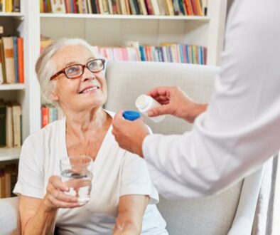 An older woman with glasses holds a glass of water as someone offers her a pill bottle—Medicare enrollment support at home.