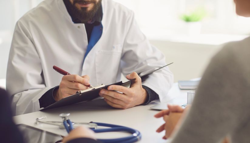 A doctor holding a clipboard and pen explains Medicare terms with a patient; a stethoscope lies on the desk.