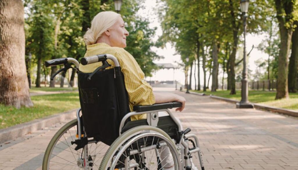 An older woman in a wheelchair sits on a paved path in a park, surrounded by trees and greenery.