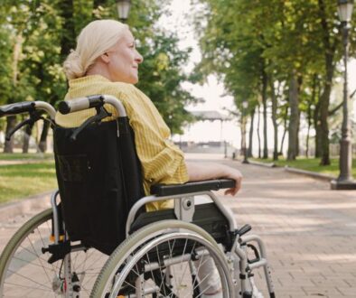 An older woman in a wheelchair sits on a paved path in a park, surrounded by trees and greenery.