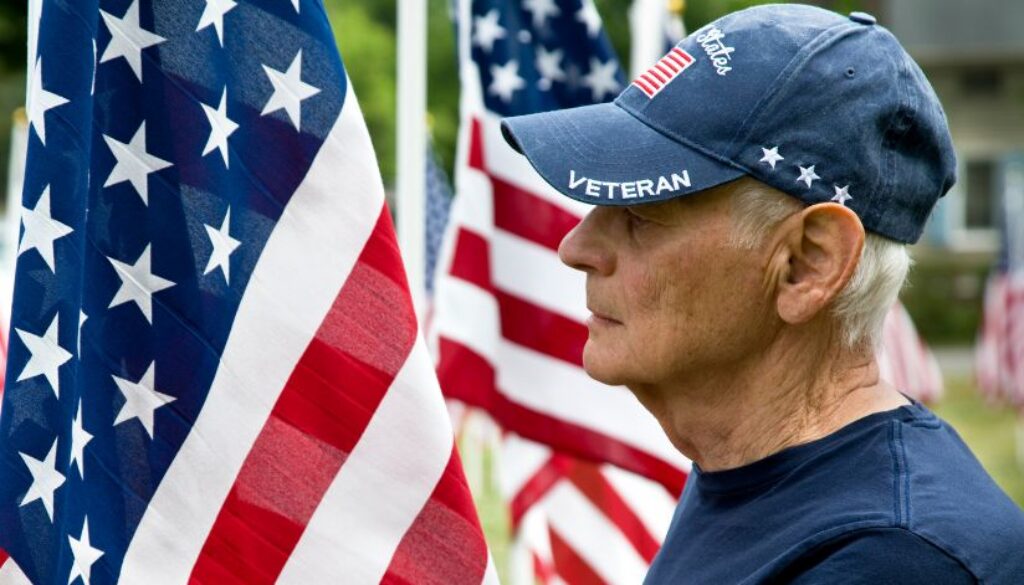 Older man wearing a “Veteran” cap stands among several American flags, looking to the right.