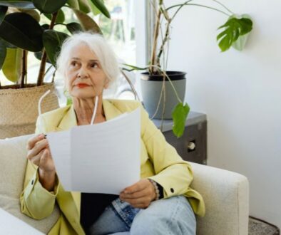 Older woman in a yellow blazer sits on a couch holding papers, looking to the side near indoor plants.