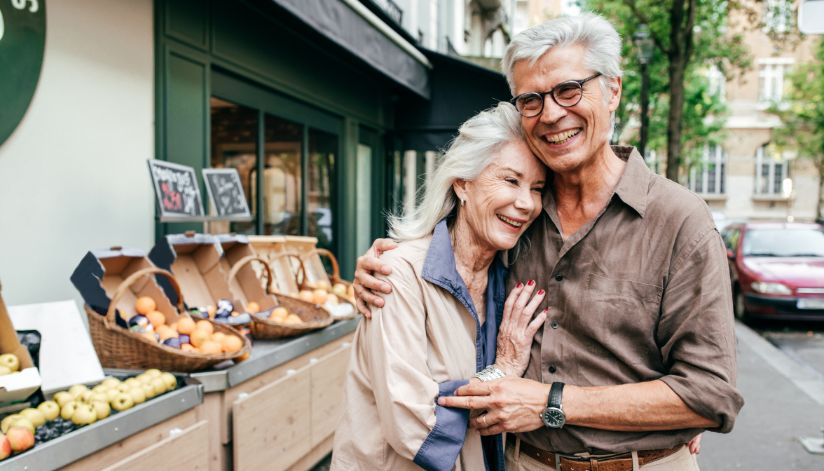 Older couple smiling and hugging while standing in front of a fruit stand on a city street after factoring healthcare in their retirement planning.