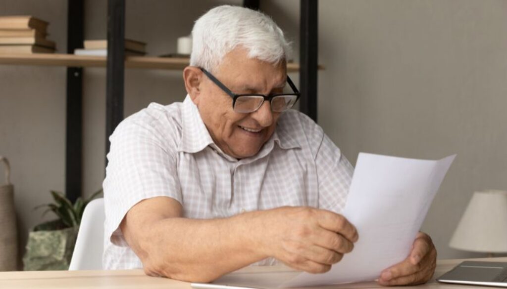 Older man with glasses sits at a desk, smiling while reading a piece of paper in a home office setting.