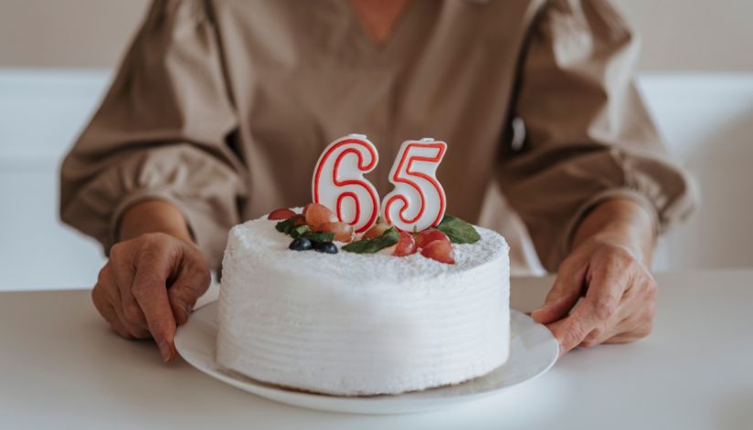 A person holds a white frosted cake with “65” candles and fruit decorations on top, sitting at a white table.