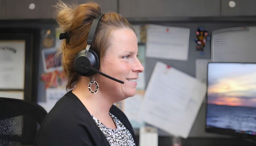Woman wearing a headset sits at a desk, looking at a computer monitor with documents pinned on the wall behind her reviews questions to ask a Medicare agent.
