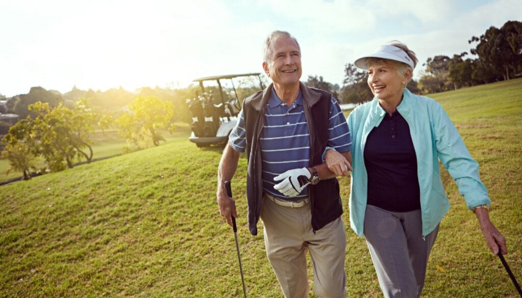 An older couple, in their first year on Medicare, walk arm in arm with golf clubs on a sunny golf course, smiling.