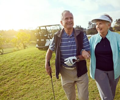 An older couple, in their first year on Medicare, walk arm in arm with golf clubs on a sunny golf course, smiling.